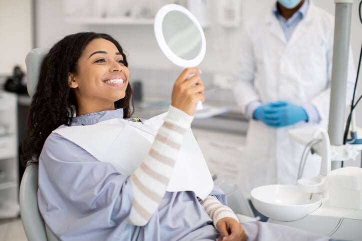 A woman in a dentist chair admiring her smile in a hand mirror