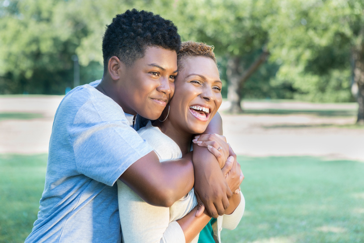 Cheerful African American teenage son gives his mom a hug from behind.