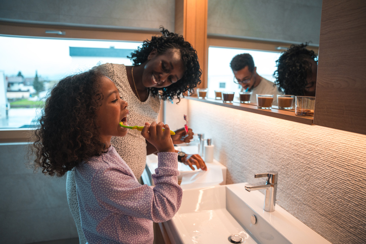 Caring African American female teaching her daughter to brush her teeth. They are the bathroom wearing pajamas.