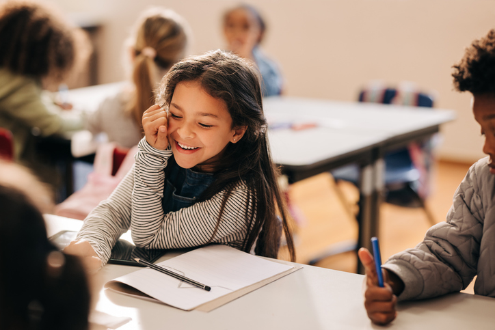 Small girl laughs happily in a primary school class, she is sitting at a table with her her classmates. Elementary school students enjoying an art and creativity class in a child development center.