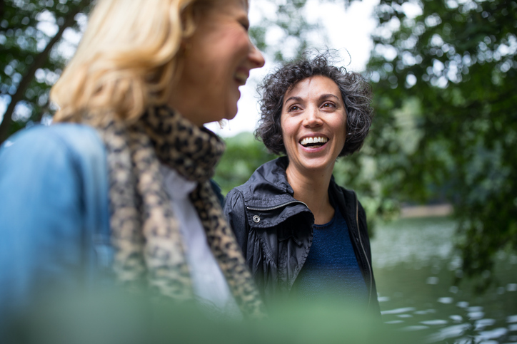 Happy mature woman looking at friend in forest. Female friends are sitting at lakeshore. They are communicating in woodland.