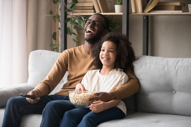 Cheerful african american father and child girl laughing holding snack remote control enjoying television comedy movie, happy black family dad with kid girl watching funny tv show sitting on sofa