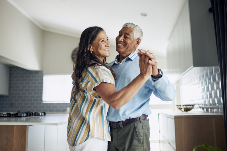 A happy elderly couple is dancing in the kitchen. They are smiling and holding each other close.