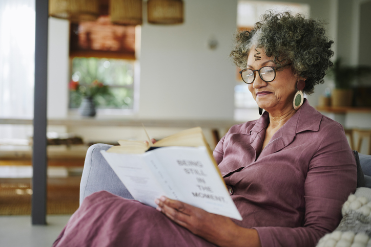 Mature woman reading a book while sitting on a sofa in her living room at home and in the afternoon