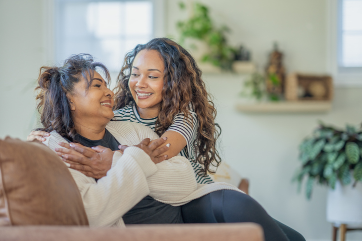 A young woman sits with her Mother on the sofa as she embraces her warmly. They are both dressed casually and are smiling as they spend time together at home.