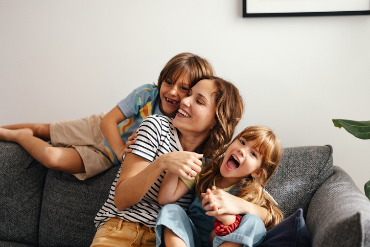 Close up shot of a young happy woman cuddling with her kids on the sofa at home. Her son and daughter are smiling while being playful and looking at the camera. The mother is smiling while hugging the children.